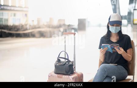 Asiatische Frau Reisende tragen chirurgische Zeichen mit Gepäck auf dem internationalen Flughafen ein Buch zu lesen, während sie auf ihren Flug warten, während der Covid Stockfoto