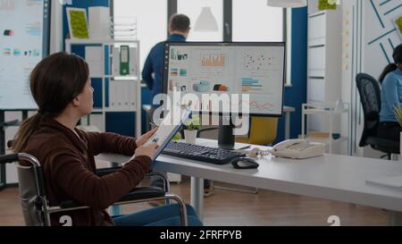 Junge Geschäftsfrau mit Behinderungen, Behinderte ungültig immobilisiert Überprüfung der Finanzberichte aus der Zwischenablage, Blick auf Computer Analyse Grafiken sitzen im Rollstuhl in Start-up-Business-Büro Stockfoto