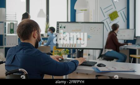 Immobilisierter Geschäftsmann mit Schutzmaske, der in einem neuen normalen Finanzunternehmen arbeitet, das am pc tippt, Berichte überprüft und Daten analysiert, die auf den im Rollstuhl sitzenden Schreibtisch blicken Stockfoto