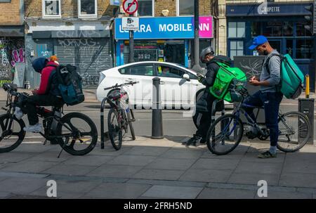 London. UK- 05.18.2021: Junge Männer arbeiten als selbstständige Reiter für Online-Lebensmittelbestellungsunternehmen, die auf ihre nächste Arbeitsbenachrichung am Telefon warten. Stockfoto