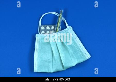 Gesichtsmaske mit medizinischem Thermometer und Pillen auf blauem Hintergrund. Schutz und Behandlung während einer Influenza- und Coronavirus-Pandemie. Stockfoto