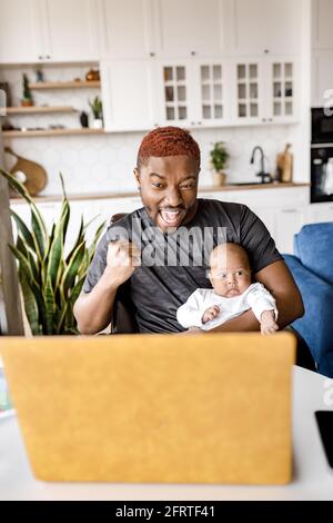 Überglücklich afroamerikanischer Arbeitsvater mit Baby-Sohn, der auf den Laptop-Bildschirm schaut und im Büro erfolgreiche und unglaubliche Online-Nachrichten feiert. Emotional Stockfoto