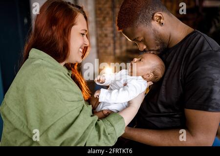 Glückliche multirassische gemischte Familie schön halten neugeborenen Sohn. Rothaarige kaukasische Mutter, die ihr Kind ansieht, afroamerikanischer Vater küsst sich sanft Stockfoto