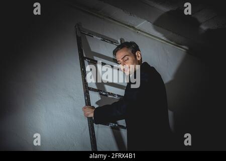 Ein junger erwachsener Mann steht mit geschlossenen Augen, meditiert oder denkt, während er sich greift, auf einer Leiter sitzt, Spanien. Er ist lässig in Schwarz gekleidet Stockfoto