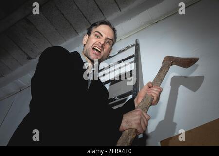 Ein psychopathischer Verrückter, junger Erwachsener, droht mit einer alten und rostigen Axt in seinen Händen, während er auf einer Leiter sitzt und bereit ist, mit einer Gambe anzugreifen Stockfoto