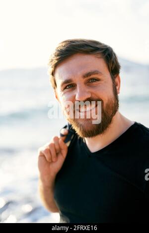 Lächelnder Mann in einer Jeansjacke über der Schulter steht vor dem Hintergrund des Meeres. Nahaufnahme. Hochformat Stockfoto