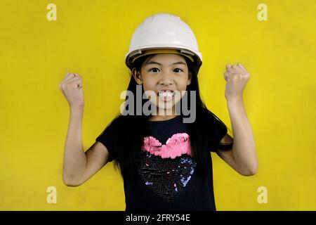 Eine Halbkörperaufnahme eines niedlichen jungen asiatischen Mädchens, das einen weißen Hut oder einen Bauhelm trägt und sich auf eine Baustelle vorbereitet. Stockfoto