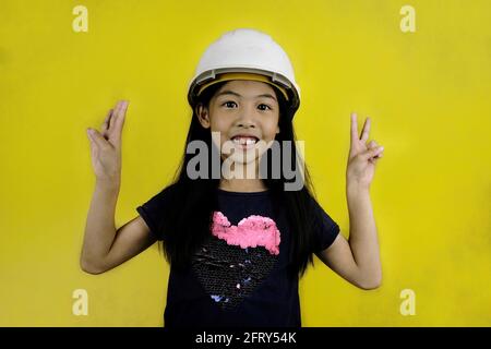 Eine Halbkörperaufnahme eines niedlichen jungen asiatischen Mädchens, das einen weißen Hut oder einen Bauhelm trägt und sich auf eine Baustelle vorbereitet. Stockfoto