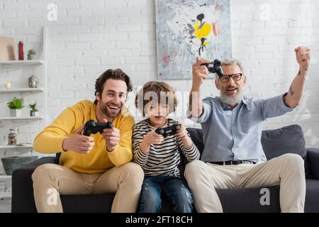 KIEW, UKRAINE - 12. APRIL 2021: Reifer Mann mit Ja-Geste in der Nähe von Sohn und Enkel mit Joysticks Stockfoto