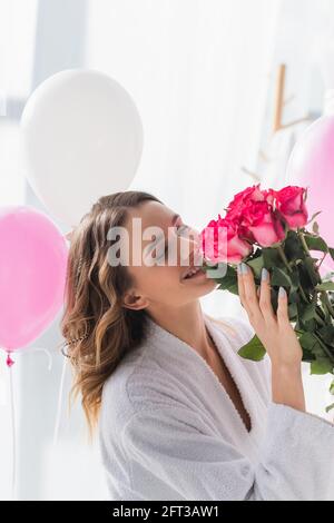 Fröhliche Frau im Bademantel riechende Rosen in der Nähe von Ballons während des Geburtstages Feiern Stockfoto