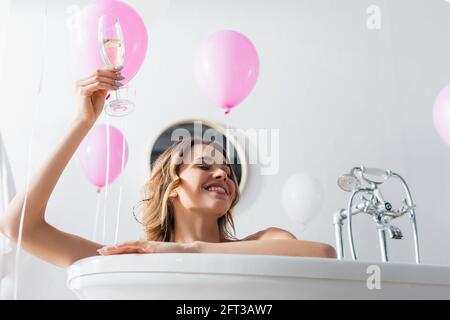 Blick aus der unteren Perspektive auf eine lächelnde Frau mit einem Glas Champagner Sitzen in der Badewanne in der Nähe von festlichen Luftballons Stockfoto