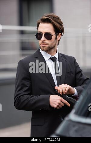 Bärtiger Bodyguard in einer Sonnenbrille, der die Waffe in der Nähe des verschwommenen Autos hält Stockfoto