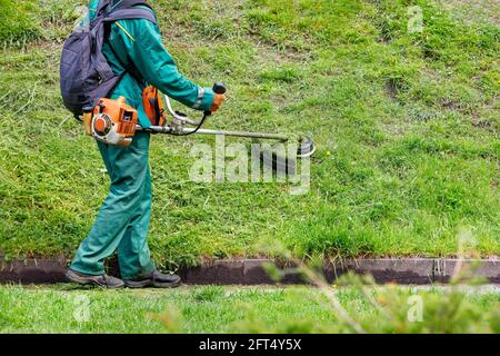 Ein Arbeiter in grünen Overalls mäht das Gras mit einem industriellen Benzintrimmer in einem Stadtpark. Speicherplatz kopieren. Stockfoto