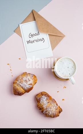 Flache Lay-Komposition mit Tasse Kaffee, Croissants und Postkarte mit Text Guten Morgen auf rosa grauem Hintergrund. Frühstückskonzept am Morgen. Draufsicht. Stockfoto