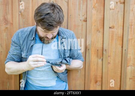 Konzentrierter Mann reinigt die Gläser vorsichtig vom Staub und wischt sie mit einem Hemd vor dem Hintergrund einer Holzwand ab. Speicherplatz kopieren. Stockfoto