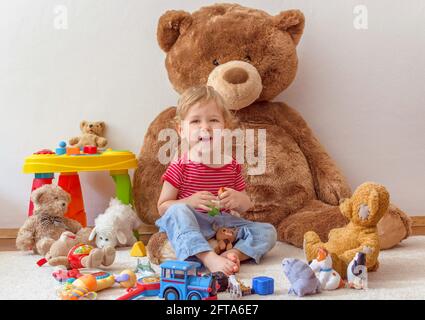 Süßer glücklicher Junge, der Spaß mit seinem riesigen Teddybären und vielen bunten Spielzeugen hat, drinnen zu Hause Stockfoto