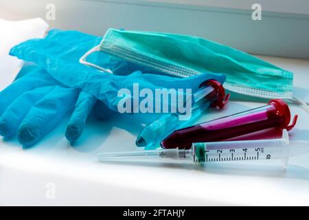 Zentrifugenröhrchen mit blauer und roter Substanz ähnlich einem Medikament oder Impfstoff. Medizinischer Handschuh, Spritze und Maske in der Nähe von Reagenzgläsern. Themenfoto: Gesundheit und Stockfoto