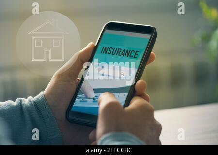 Nahaufnahme eines jungen kaukasischen Mannes, der an einem Tisch in seinem Wohnzimmer sitzt, mit einem simulierten Suchvorgang online eine Hausversicherung mit seinem Smartphone durchsucht Stockfoto