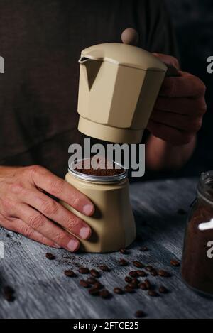 Ein junger Kaukasusmann bereitet Kaffee in einer beigen Mokakapfanne zu, die an einem grauen rustikalen Holztisch sitzt Stockfoto