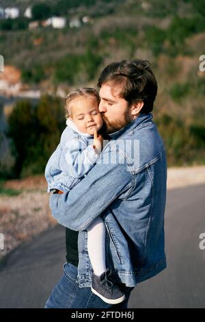 Vater trägt und küsst ein kleines Mädchen in seinen Armen, das auf der Straße steht. Nahaufnahme Stockfoto