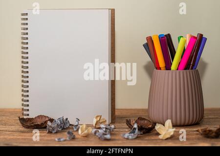 Offenes Spiral-Notizbuch mit weißem Blatt, farbigen Markern im Glas daneben und Dekorationen auf dem Tisch Stockfoto