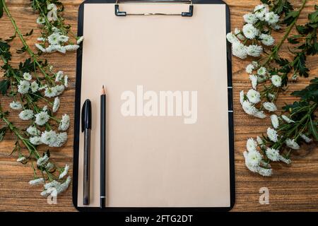 Schwarzes Klemmbrett mit Elfenbeinblatt, darauf ein Stift und ein Bleistift. Auf beiden Seiten der Zwischenablage befinden sich weiße Blumen. Stockfoto