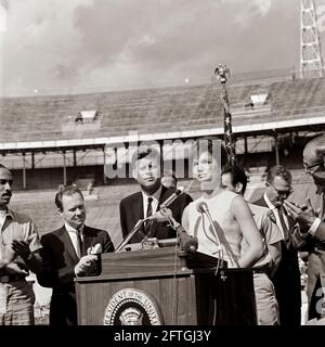 First Lady Jacqueline Kennedy hält bei einer Zeremonie der 2506. Flagge der kubanischen Invasionsbrigade im Orange Bowl Stadium in Miami, Florida, Bemerkungen auf Spanisch; Mitglieder der Brigade überreichten ihre Flagge an Präsident John F. Kennedy und Frau Kennedy. Auf der Rednerplattform (von links nach rechts): Erneido Oliva, zweiter Kommandant der Brigade (applaudierende); Bürgermeister von Miami, Robert King High (applaudierende); Präsident Kennedy; Jose Pérez San Román, Kommandant der Brigade (hinten, meist versteckt); die First Lady (an Rednermikrofonen); Manuel Artime, Chef der zivilen Brigade . Stockfoto