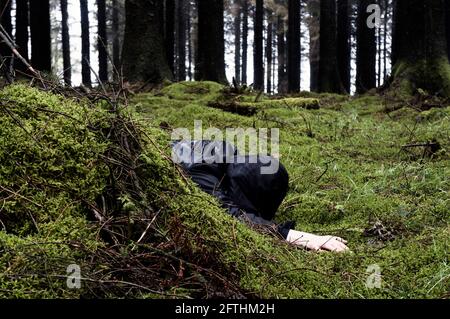 Ein Mordkonzept, das sich auf ein Verbrechen eines mit Kapuze versehenen Mannes, der an einem düsteren, nassen Tag mit dem Gesicht nach unten in einen dunklen Wald zeigt Stockfoto