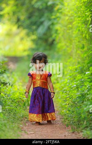 Südindische niedliche Mädchen Kind trägt schöne traditionelle Kleid langen Rock und Bluse, zu Fuß durch Dorf Weg mit viel Grün Hintergrund. Stockfoto