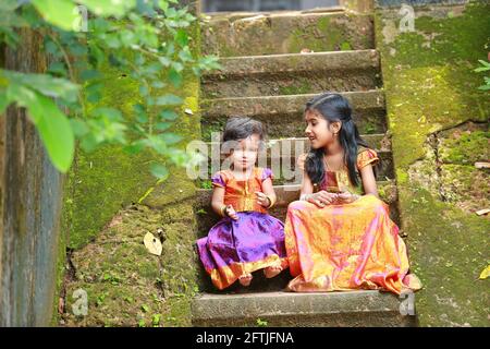 Südindische Mädchen Kinder tragen schöne traditionelle Kleid langen Rock Und Bluse, sitzend auf den langen Stufen eines bedeckten Hauses Mit moosgrünem Pilz Stockfoto