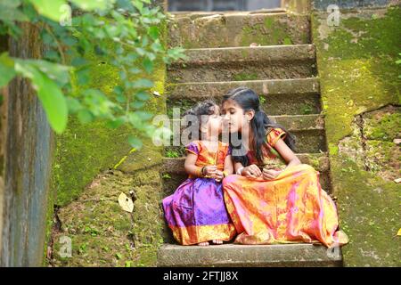 Südindische Mädchen Kinder tragen schöne traditionelle Kleid langen Rock Und Bluse, sitzend auf den langen Stufen eines bedeckten Hauses Mit moosgrünem Pilz Stockfoto