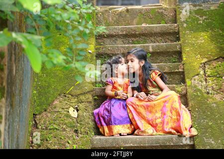 Südindische Mädchen Kinder tragen schöne traditionelle Kleid langen Rock Und Bluse, sitzend auf den langen Stufen eines bedeckten Hauses Mit moosgrünem Pilz Stockfoto