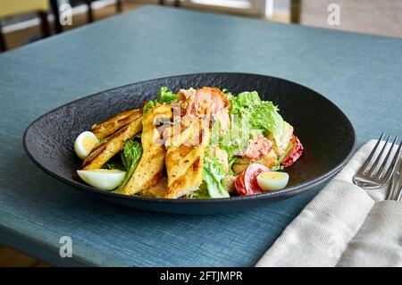 Frischer Caesar-Salat mit Huhn in einer schwarzen Schüssel auf der Veranda des Restaurants. Stockfoto