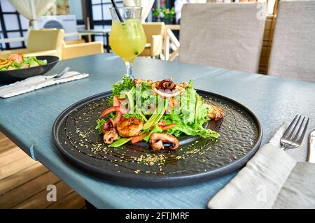 Frischer mediterraner Salat mit Garnelen, Tintenfisch und Tintenfisch auf einem schwarzen Teller auf der Veranda des Restaurants. Stockfoto