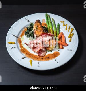 Ein Gericht aus Seebarsch-Filetsteak mit Speck, Spinat, Spargel und Baby-Karotten Stockfoto