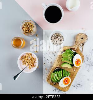 Gesundes Frühstückskonzept. Sandwich mit Avocado und gekochten Eiern, Joghurt mit Müsli, Tasse Kaffee auf tricolor stilvollem Hintergrund, Draufsicht, flach Stockfoto