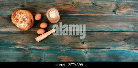 Frisch gebackener ganzer Laib Sauerteig, Mehl, Eier und Nudelholz auf verblassten blauen Holzplanken im flachen Lay-Format Stockfoto