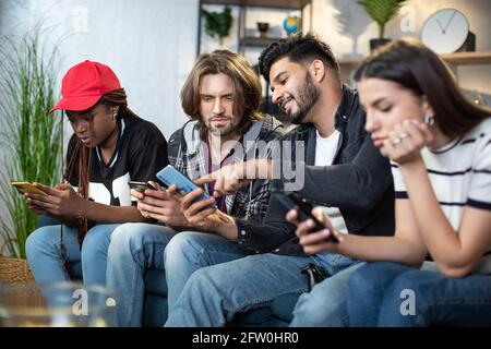 Männer und Frauen entspannen sich auf der bequemen Couch mit Gadgets in den Händen. Vier gemischte Rennfreunde treffen sich zusammen, um Freizeit online zu verbringen. Moderne Lebensstile junger Menschen. Stockfoto