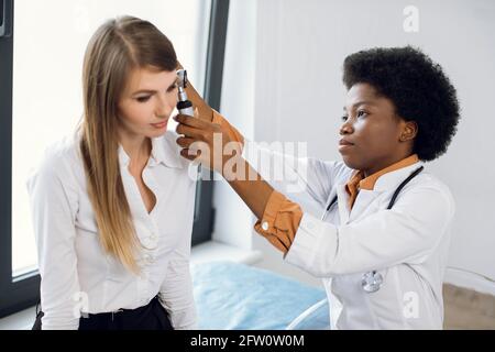 HNO-Arzt und Patient im modernen Krankenhaus. Weibliche selbstbewusste afroamerikanische Ärztin, die Ohrenuntersuchung mit Otoskop für ihre Patientin, junge europäische Dame, die an Ohrenschmerzen und Otitis leidet, macht Stockfoto