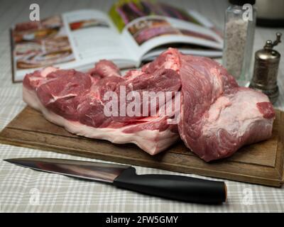 Ungekochte Schweinefleisch auf Holzbrett mit Messer, Salzmühle, Pfeffermühle und Rezeptbuch. Frisches, rohes Schweinefilet Stockfoto