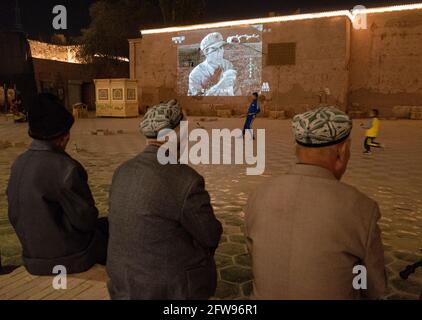 Uyhgur Männer sitzen auf einem kleinen Platz und schauen sich einen Propagandafilm über die Chinesische Revolution an. Kashgar , Volksrepublik China 2019 Stockfoto