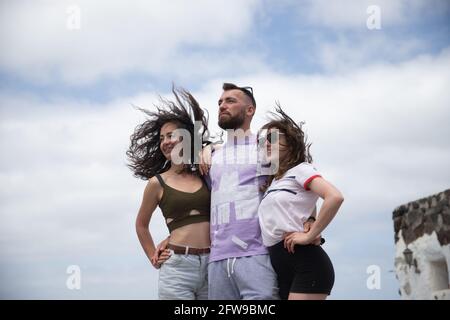 Eine Gruppe von Freunden, zwei Frauen und ein Mann, umarmen und betrachten einen Punkt alle zusammen, sie sind isoliert mit dem Himmel als Hintergrund, Konzept der Vielfalt. Stockfoto