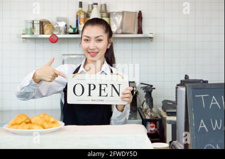 Ein junger asiatischer Ladenbesitzer hält mit einem Lächeln ein OFFENES Schild vor einem Café-Schalter. Morgendliche Atmosphäre in einem Café. Stockfoto