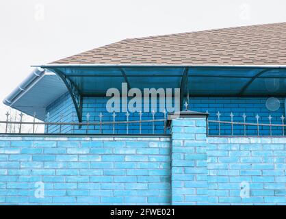 Die Zaunwand ist aus Ziegelstein in blau lackiert, das Dach des Hauses ist sichtbar von hinter dem Zaun, moderne Hütte Konstruktion Stockfoto