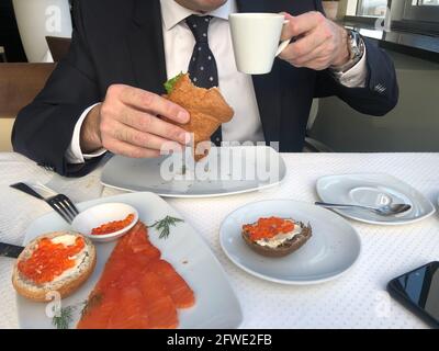 Mann in einem Business-Anzug, Krawatte und weißem Hemd frühstücken, Kaffeepause, eine Tasse Kaffee, Croissant mit Lachs und grünem Salat, Sandwiches mit Stockfoto