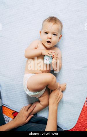 Mama hält das Baby in einer Windel mit einem Spielzeug-Igel in den Händen an den Beinen. Das Baby liegt auf einem Bettüberwurf auf einem farbigen Teppich auf dem Boden Stockfoto