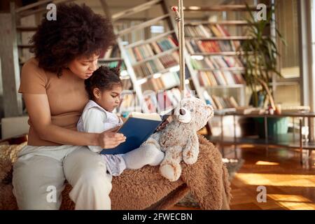 Eine junge Mutter und ihre kleine Tochter sitzen zu Hause in entspannter Atmosphäre auf der Schaukel und lesen ein Buch. Familie, gemeinsam, Freizeit Stockfoto