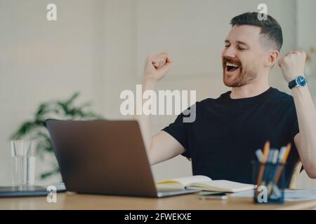 Glücklicher junger Freiberufler, der eine gewinnende Geste zeigt, während er an seinem Platz sitzt Arbeitsplatz zu Hause Stockfoto