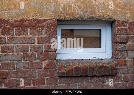 Modernes weißes Kunststofffenster mit einem Glanz, der zum Keller führt, darunter ein Ziegelregal im unteren Teil, das stellenweise mit braun abgebrochenem Material ausgekleidet ist Stockfoto