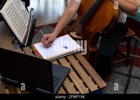Junger Musiker, der Notizen in einem Notizbuch auf einem hölzernen Tabelle in einem Online-Kurs mit Cello Laptop mit selektiven Fokus- und Kopierbereich Stockfoto